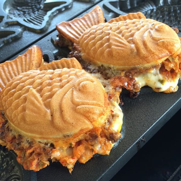 Two savory taiyaki, filled with kimchi and cheese, cooking in a fish-shaped waffle iron.