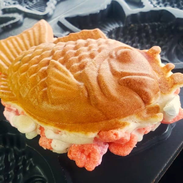 A golden-brown taiyaki, a fish-shaped waffle, filled with white and pink cream, in a dark mold.