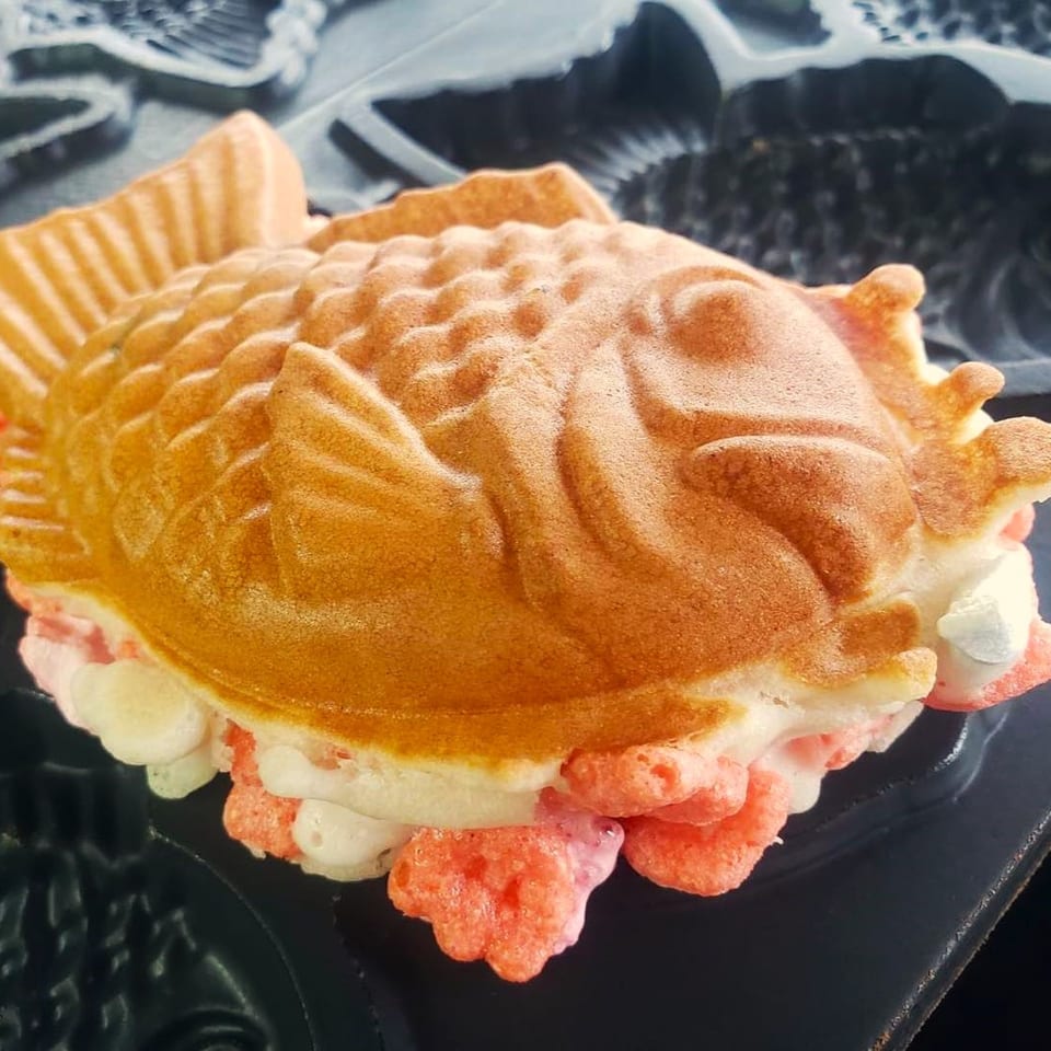 A golden-brown taiyaki, a fish-shaped waffle, filled with white and pink cream, in a dark mold.